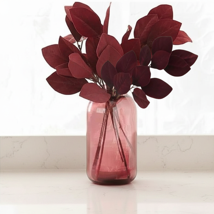 Burgundy velvet leaf sprays in a pink vase on a white surface with a light background