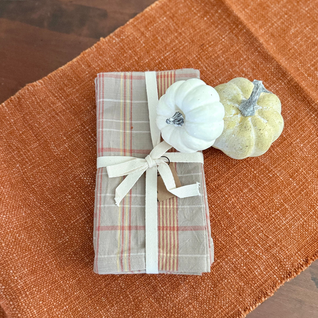 Folded plaid fabric with a white ribbon on a brown textured surface with small pumpkins.