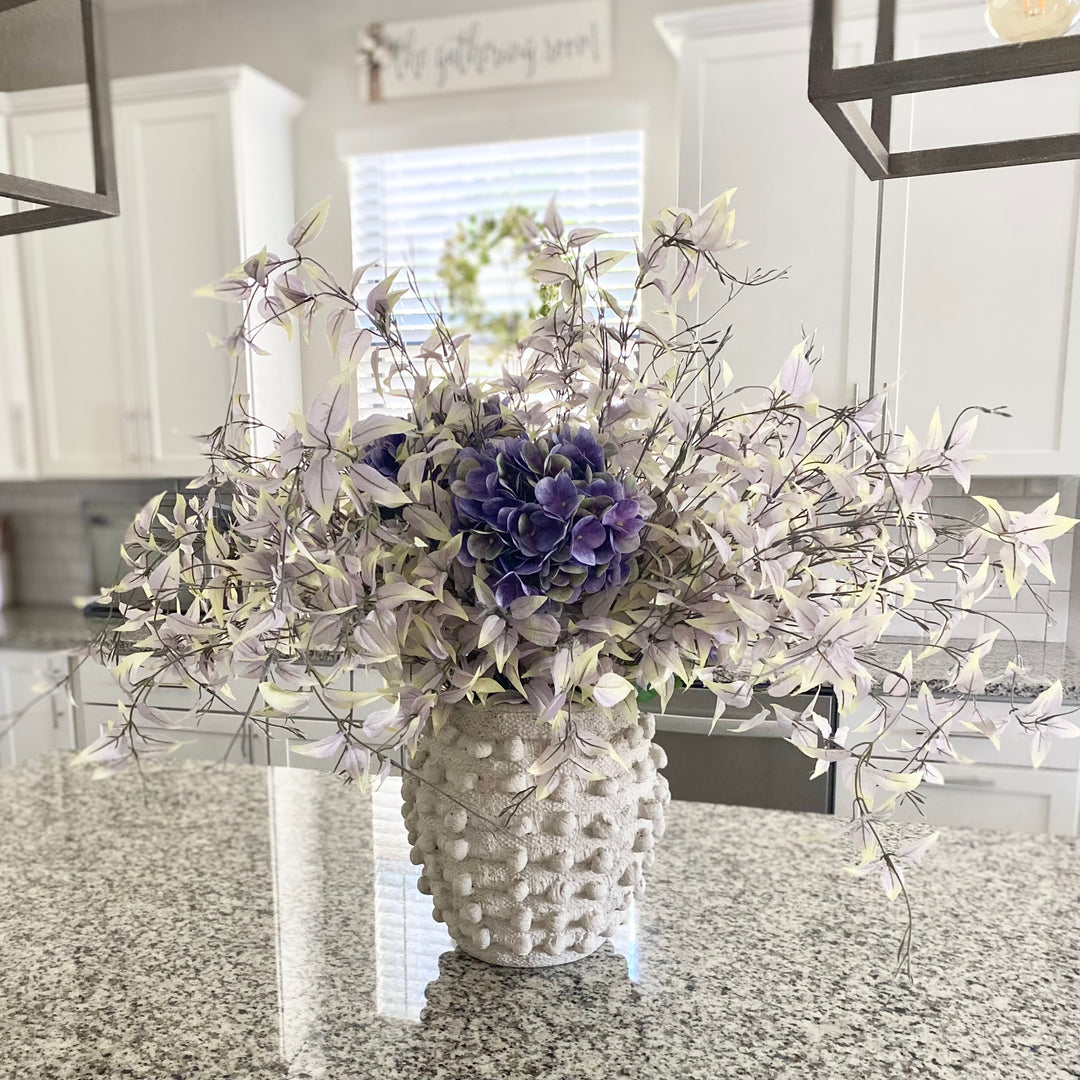 A set of dusty purple weeping leaf sprays arranged in a white vase, placed on a kitchen countertop.