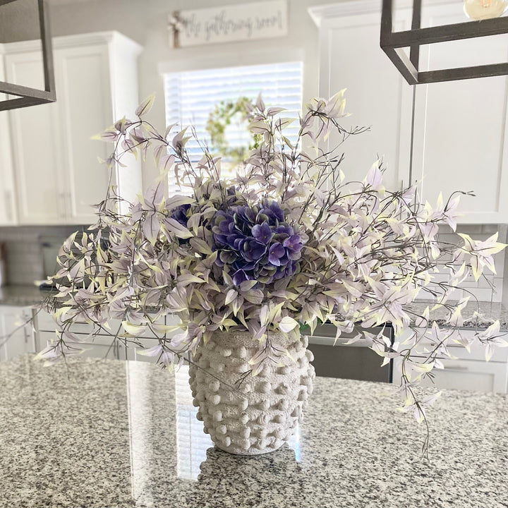 A set of dusty purple weeping leaf sprays arranged in a white vase, placed on a kitchen countertop.