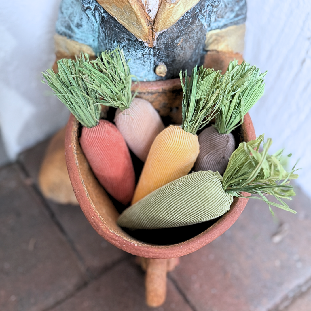 Decorative garden rabbit with a basket of colorful plush fabric carrots on a brick paver surface.