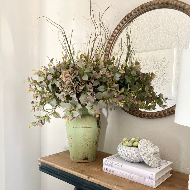 A set of four artificial eucalyptus sprays with berries in light green and purple, arranged in a tall green vase, placed on a shelf under a circular mirror.