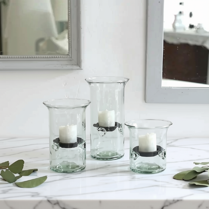 Three clear glass candle holders with black metal accents and white taper candles, arranged on a white marble surface with green leaves in the foreground and mirrors in the background.
