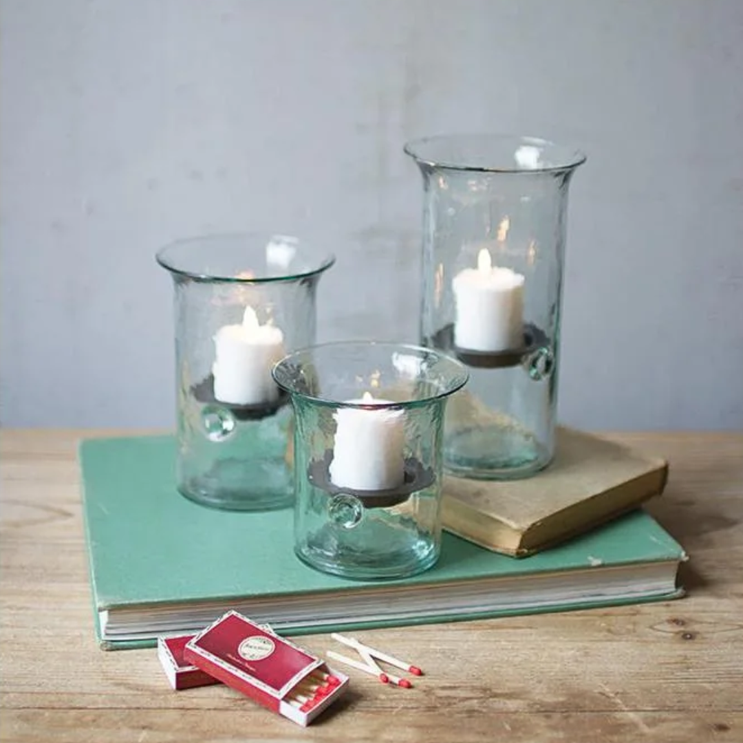 Three clear glass candle holders with white pillar candles, styled on a green book with a vintage book and matches on a wooden table against a gray wall.