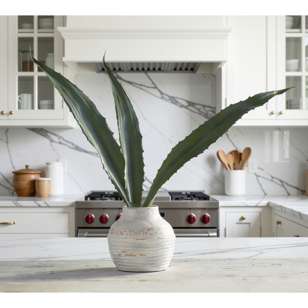 Long green faux aloe leaves in a textured white ceramic pot, set on a marble countertop in a bright, modern kitchen.