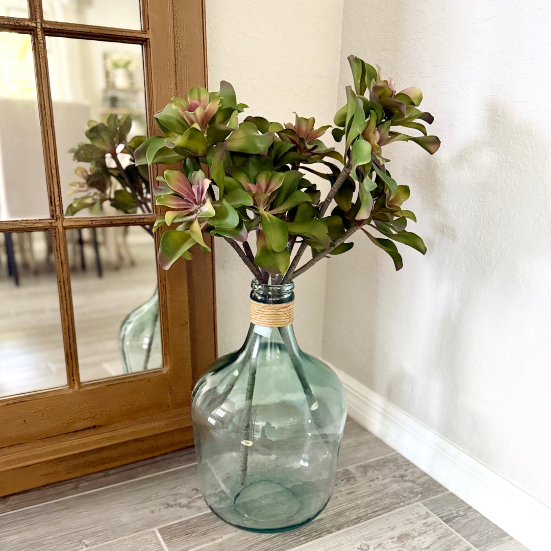 Green glass jug with artificial succulents on a gray wood plank floor next to a mirror.