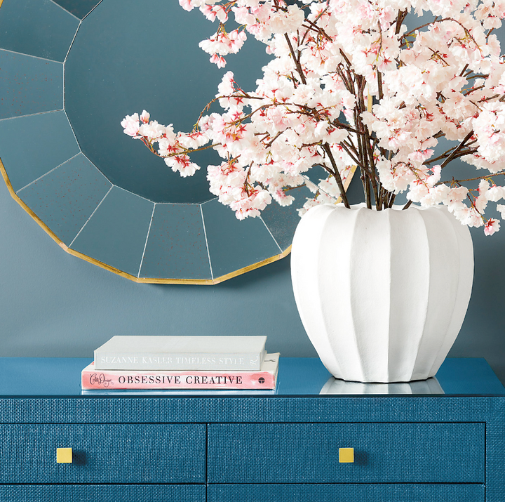White vase with pink cherry blossom branches on a blue surface with a decorative mirror in the background.