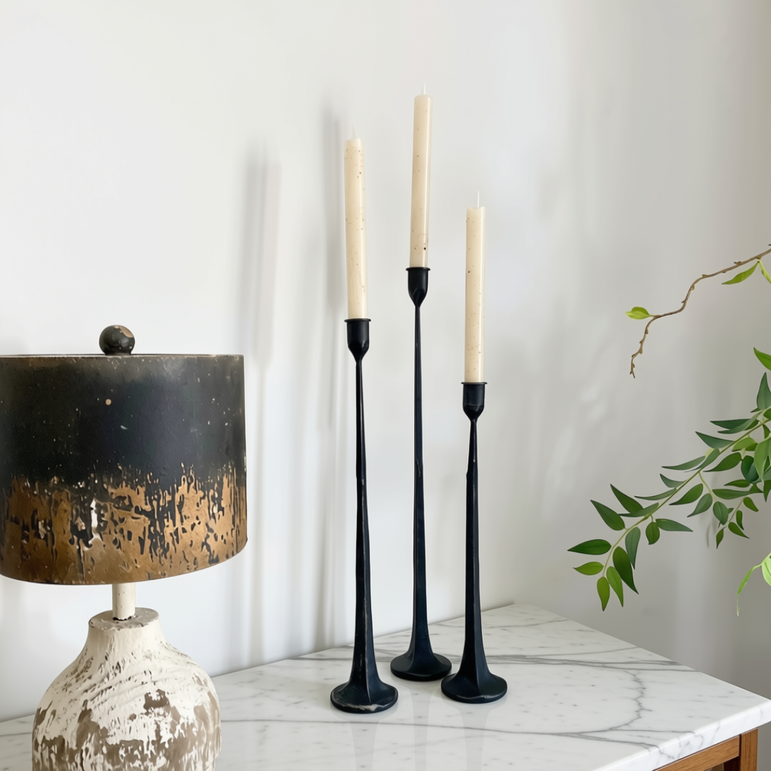 Three black cast iron taper candle holders with cream taper candles, styled on a white marble side table next to a distressed lamp and a green plant branch against a white wall.