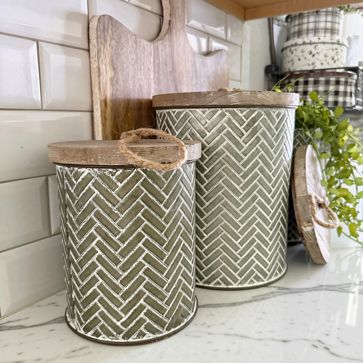 Three decorative metal canisters with wooden lids and rope handles, featuring a white herringbone pattern on a muted sage green background, styled on a white marble kitchen counter.