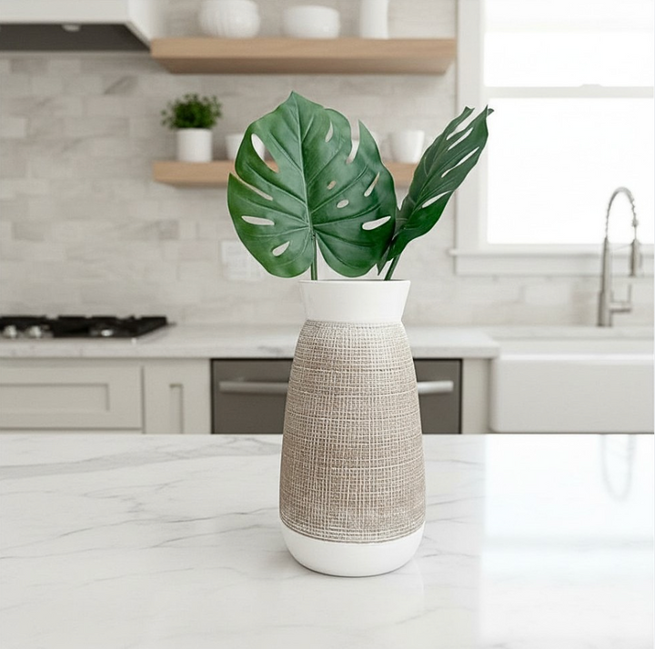 A modern white ceramic vase with a textured beige woven wrap, holding two green monstera leaves, set on a white marble kitchen countertop with a blurred kitchen background.