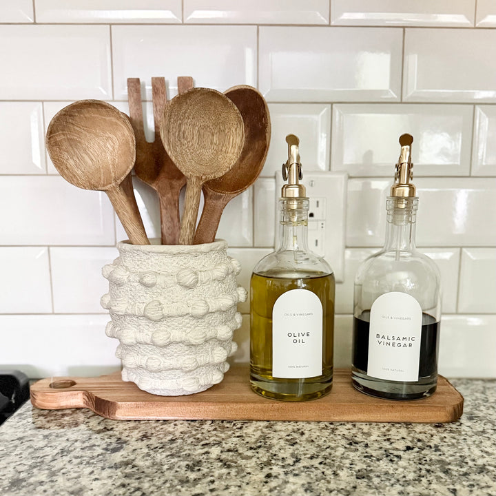 Wooden spoons in a ceramic holder with olive oil and balsamic vinegar bottles on a kitchen counter.