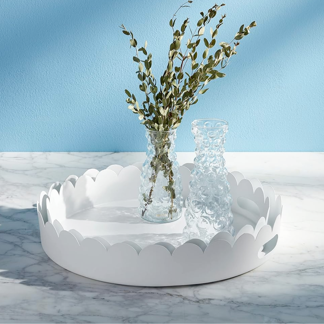 Two round white scalloped metal trays with cut-out handles, one nested inside the other, on a marble surface with a vase of greenery placed on one of the trays.