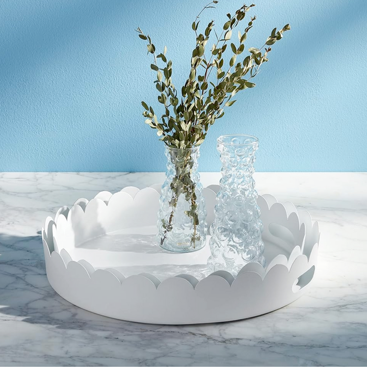 Two round white scalloped metal trays with cut-out handles, one nested inside the other, on a marble surface with a vase of greenery placed on one of the trays.