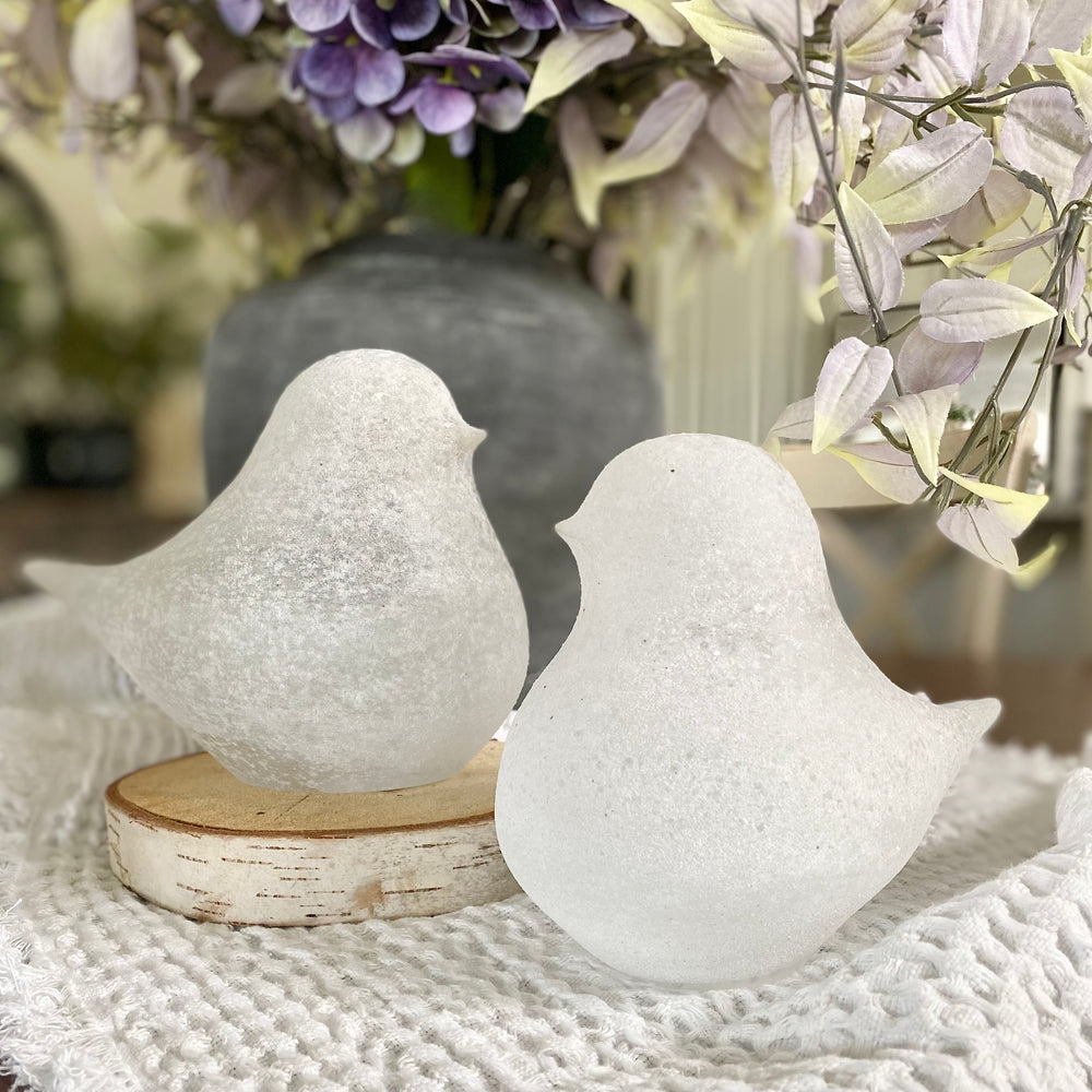 Two white frosted glass bird figurines on a wooden base, with one facing left and the other right, against a blurred background with a floral arrangement.