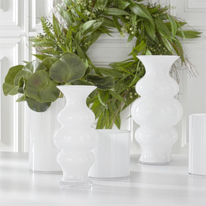 A set of white hourglass fluted glass vases in two sizes displayed on a surface with greenery in the background.