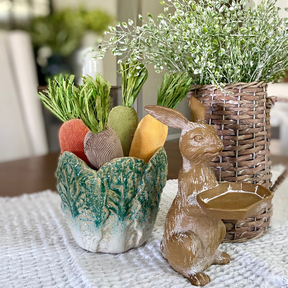 A set of plush fabric carrots in various earthy colors displayed in a home decor setting with a ceramic basket and a ceramic rabbit.