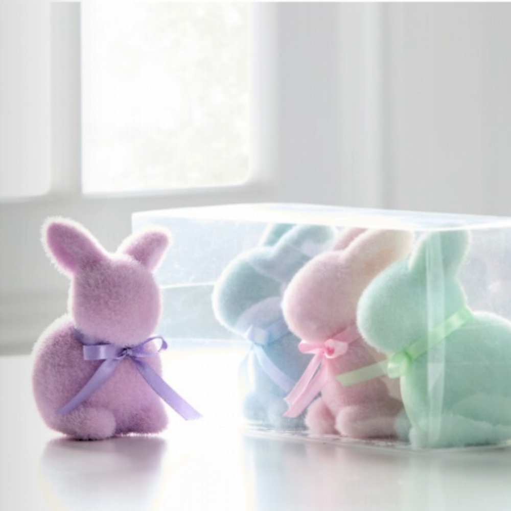 A set of four fuzzy flocked bunnies in pastel colors standing in a clear box, with one bunny wearing a purple bow.