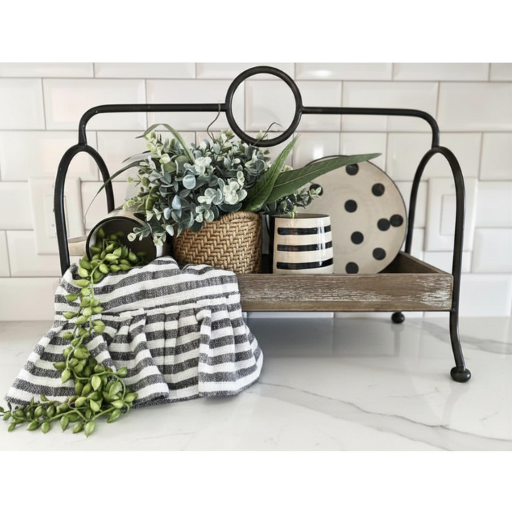 A rustic black metal bench-style tray table with a weathered wood shelf, styled with a striped throw blanket, potted greenery, a woven basket, and a ceramic plate on a white marble surface against a white subway tile background.
