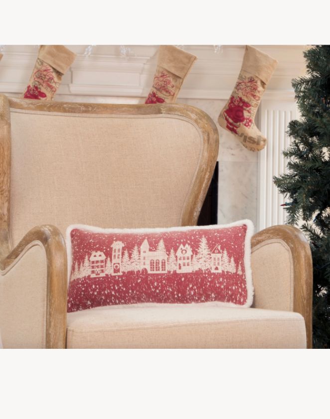 Decorative pillow with a Christmas village design on a beige armchair with stockings and a tree in the background.