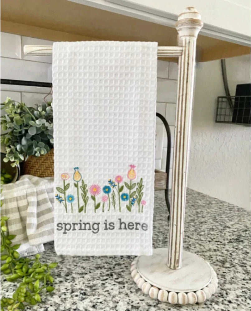 Kitchen towel with 'spring is here' text and floral design hanging on a towel rack.