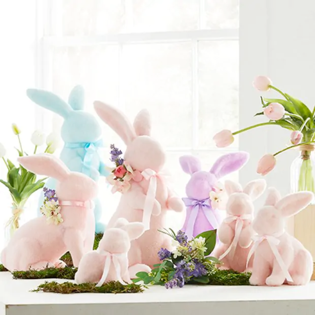 A collection of pastel pink flocked bunnies in various sizes, displayed on a surface with a floral arrangement and a window in the background.