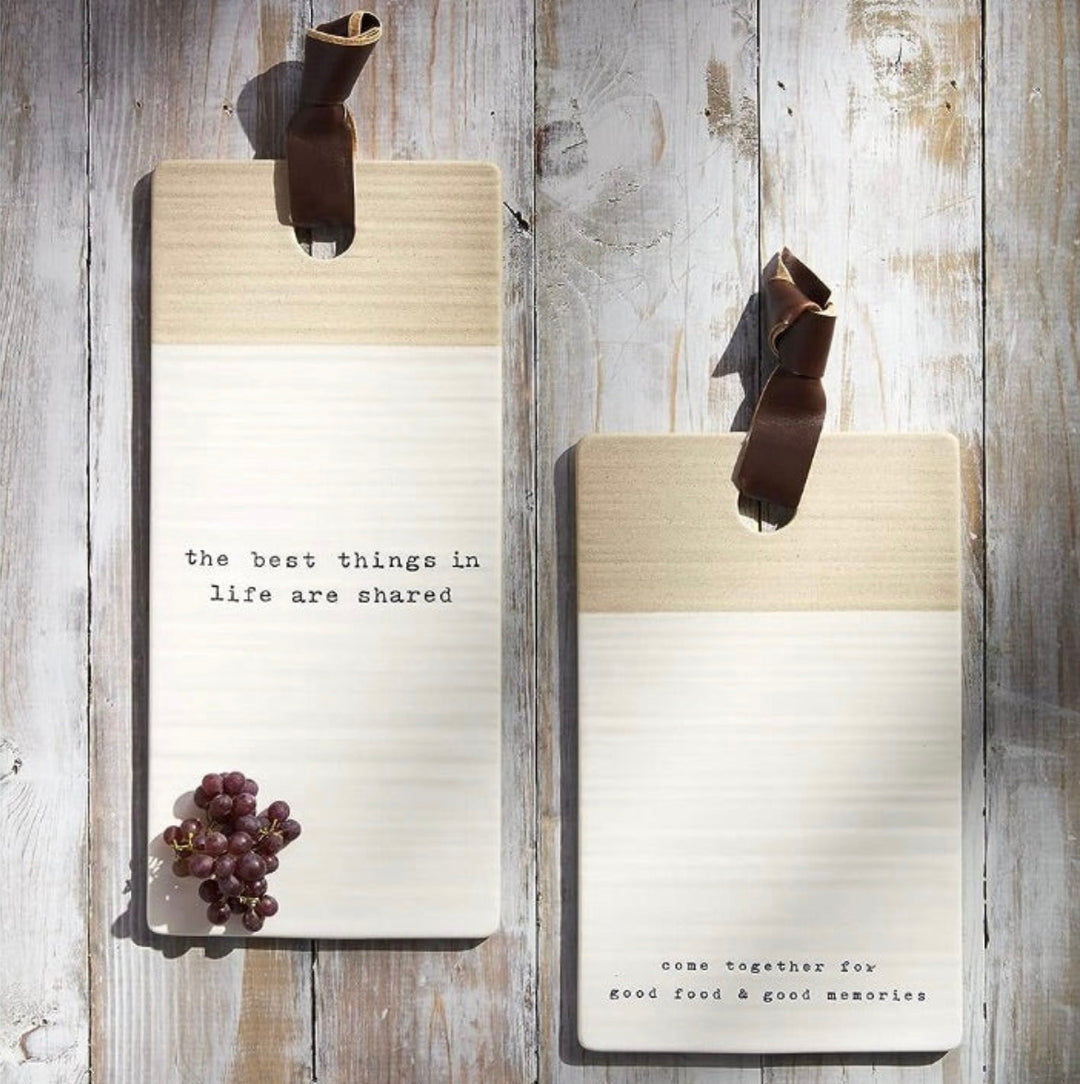 Two stoneware cutting boards, one short with the text 'come together for good food & good memories' and one long with the text 'the best things in life are shared'. Both have a faux leather loop attached to the top edge.