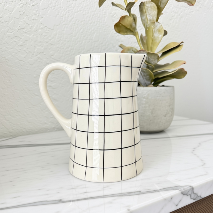 A white stoneware pitcher with a black grid pattern, sitting on a white marble countertop next to a potted succulent plant.