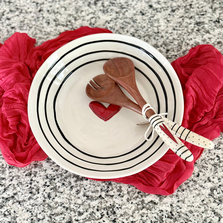 Stoneware serving bowl with a red heart and black stripes and wooden utensils on a red table runner