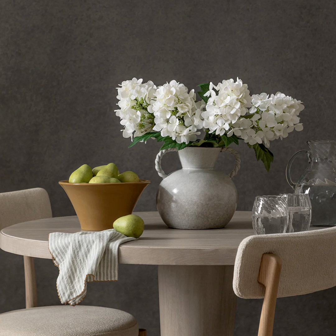 Dining table with a white stoneware vase with twisted handles. White flowers, bowl of pears, and glasses on a dark background