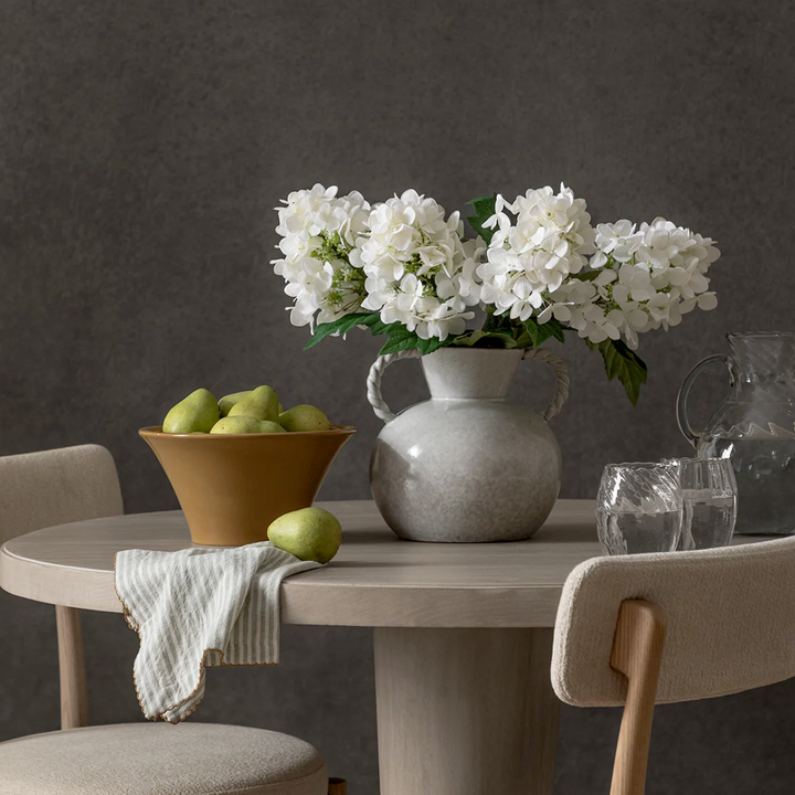 Dining table with a white stoneware vase with twisted handles. White flowers, bowl of pears, and glasses on a dark background