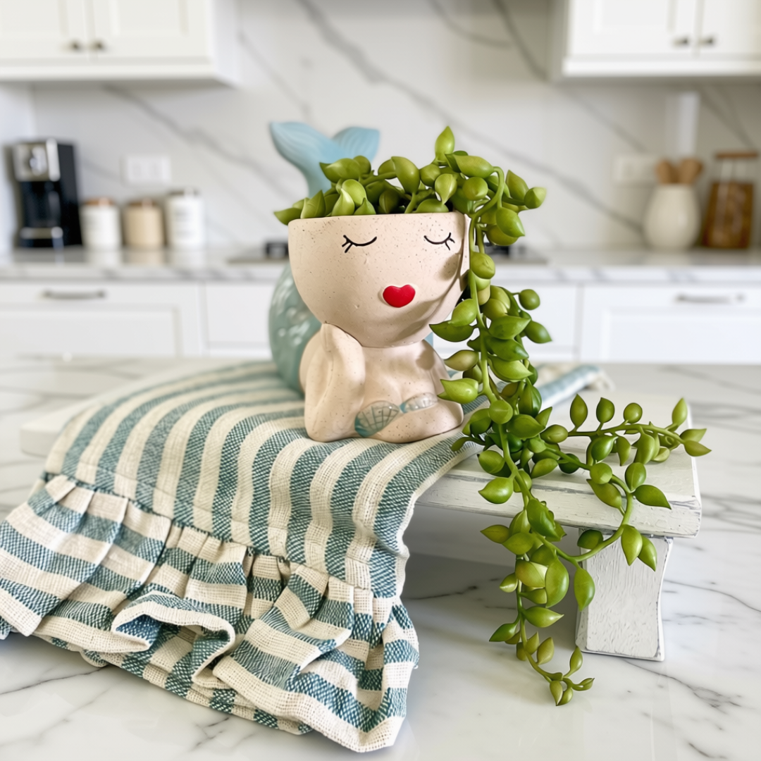 A ceramic planter shaped like a smiling mermaid with a blue tail, holding a trailing green plant, styled on a striped blue and white towel in a bright kitchen setting.