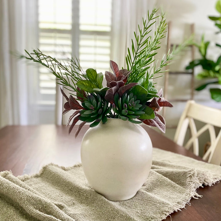 A faux succulent and mixed foliage bundle arranged in a white vase, placed on a beige fabric surface with a window in the background.