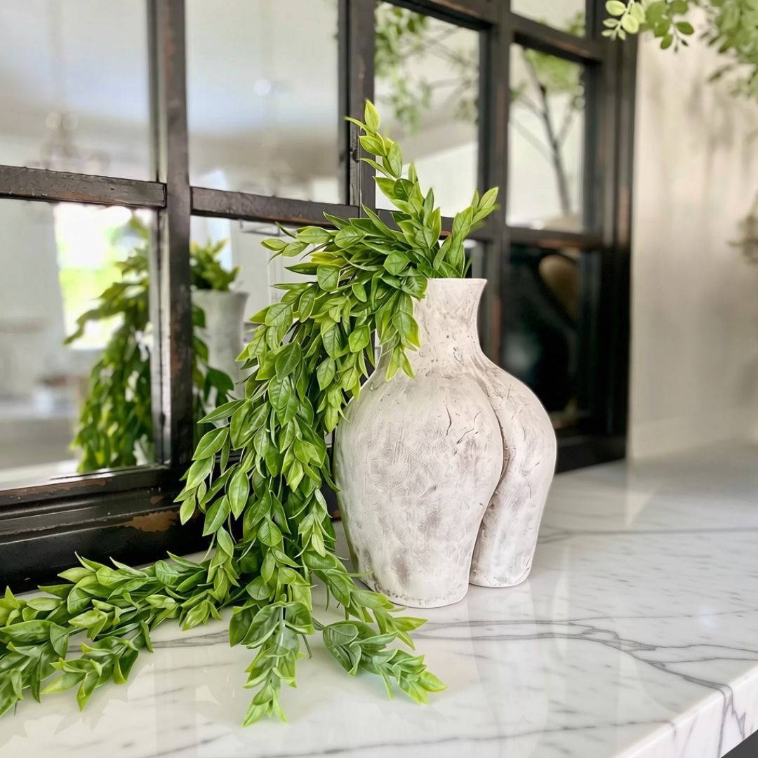 Textured clay Culo pot in off-white  styled with green leafy branches on a white marble surface in front of a black-framed window.
