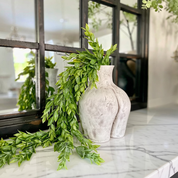 Textured clay Culo pot in off-white  styled with green leafy branches on a white marble surface in front of a black-framed window.