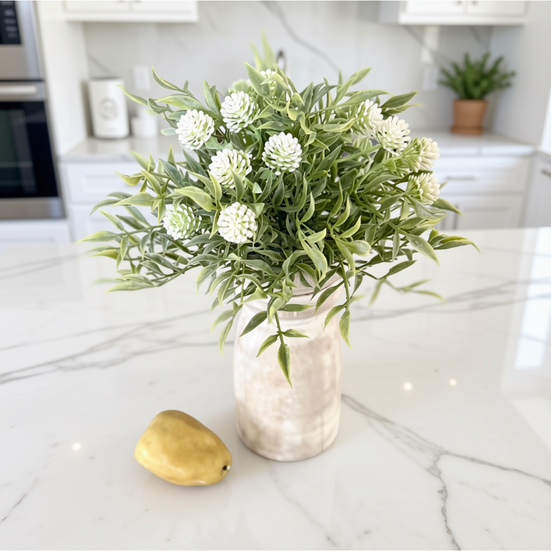 A decorative arrangement of faux green leaves and white globe flowers in a textured cream vase, placed on a white marble kitchen countertop with a small gold pear beside it.