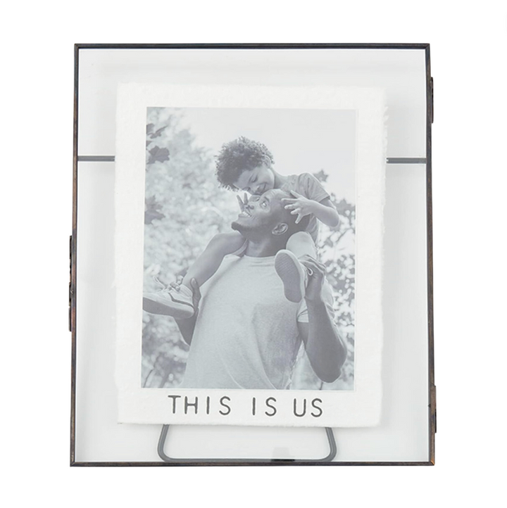 A metal picture frame with glass panes displaying a black and white photo of a person holding a young child, with the text 'THIS IS US' on the matting.
