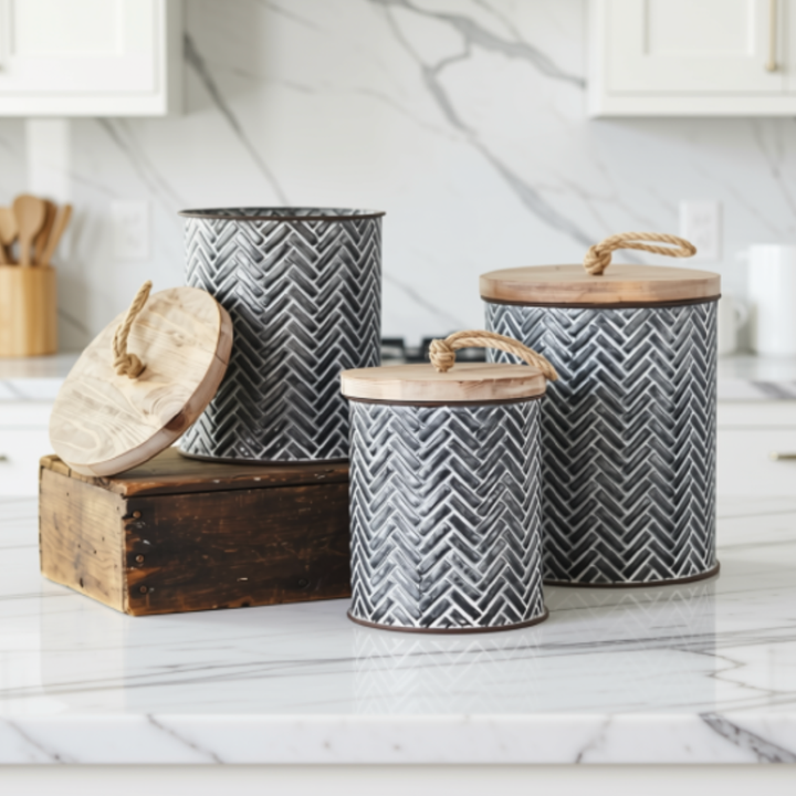 Three metal kitchen canisters with a gray and white herringbone pattern and light wood lids, displayed on a white marble kitchen counter.