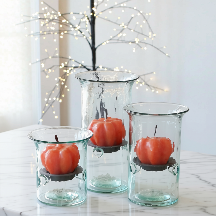Three clear glass candle holders of different sizes on a white marble surface, each holding an orange pumpkin-shaped candle, with a decorative branch of warm white string lights blurred in the background.