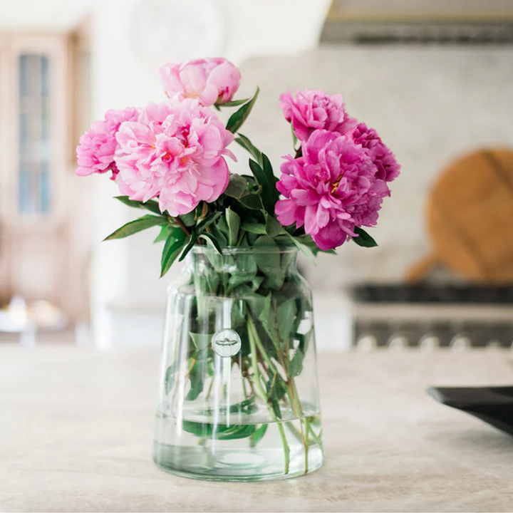 A clear glass mason jar vase with sloping sides, containing a bouquet of pink flowers, displayed on a kitchen countertop.