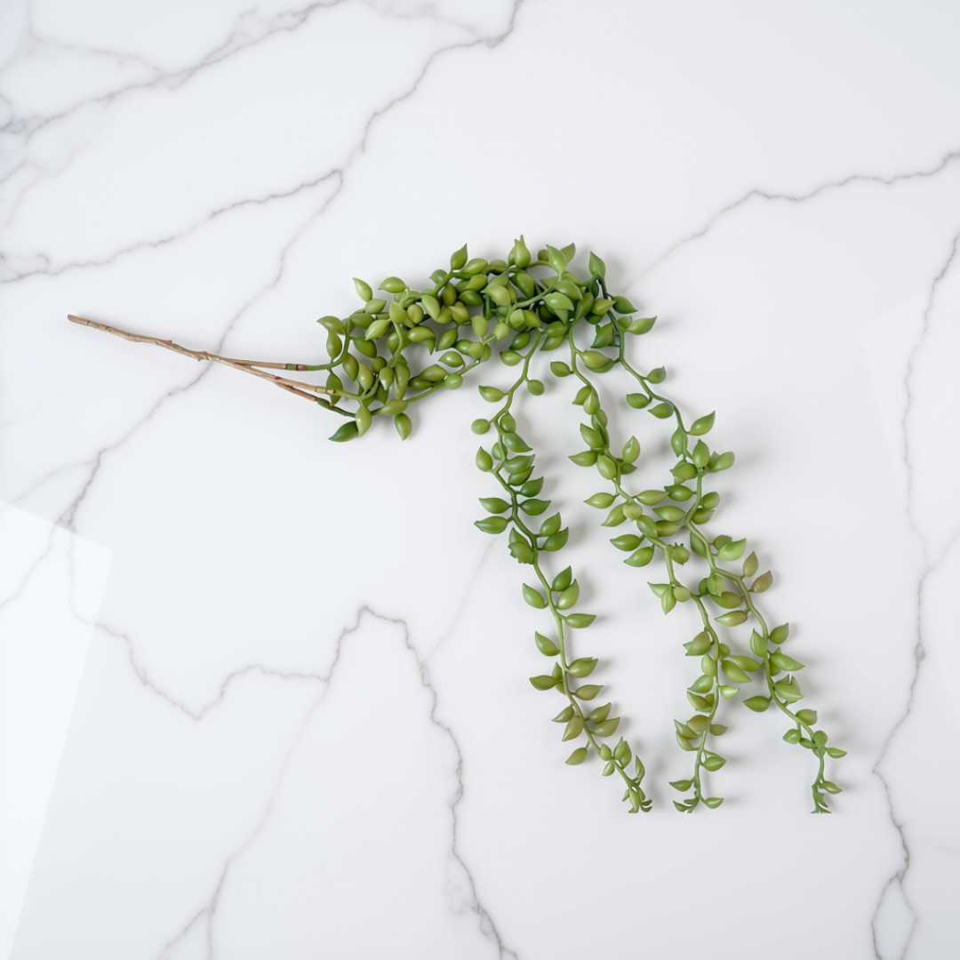 A green trailing artificial plant stem with small oval leaves and a brown twig-like base, laid on a white marble surface.