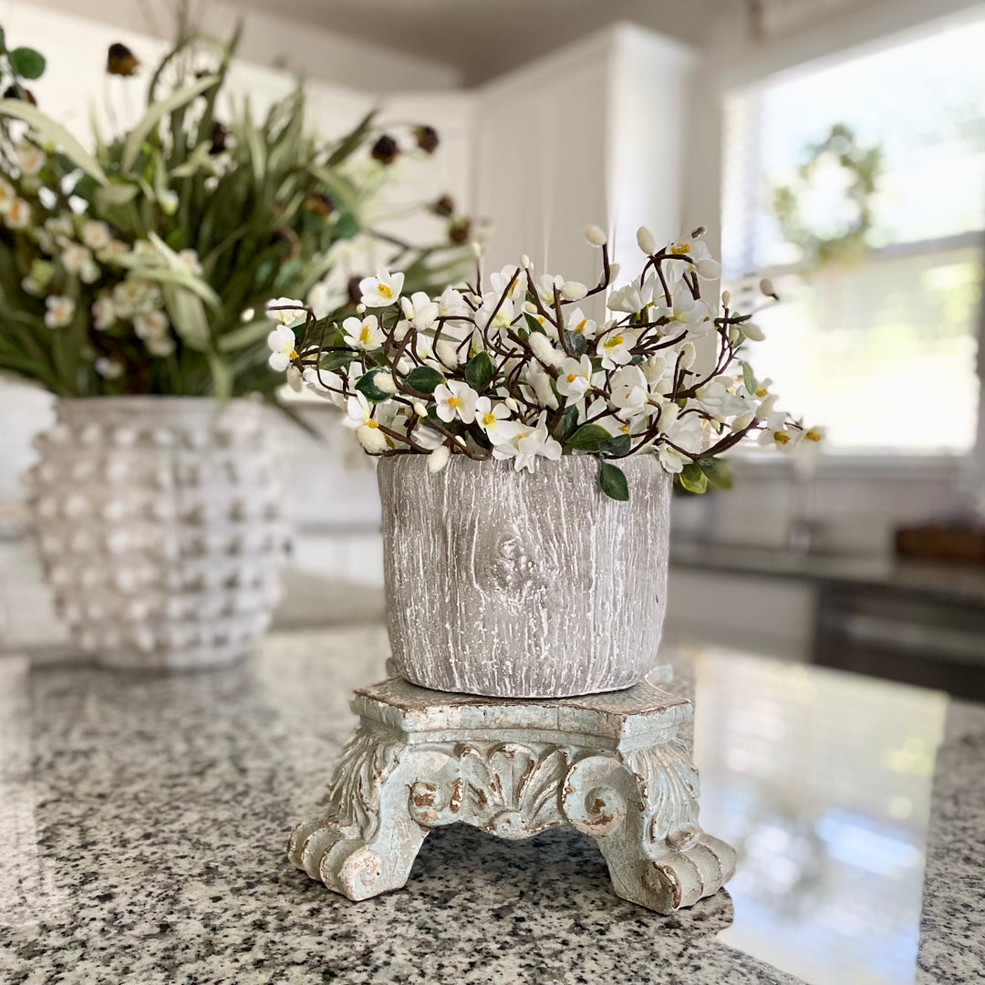 A faux Tree Bark Planter made from cement, with a textured bark finish in shades of gray. It is displayed with a arrangement of white flowers inside it, placed on a kitchen counter.