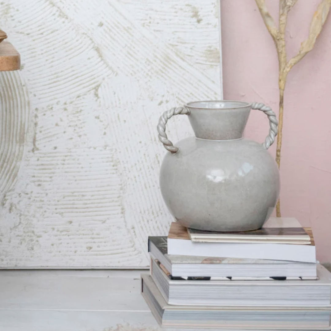 A stoneware vase with twisted handles placed on top of a stack of books, featuring a soft cream color with a reactive glaze finish.