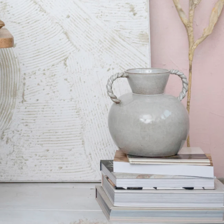 A stoneware vase with twisted handles placed on top of a stack of books, featuring a soft cream color with a reactive glaze finish.