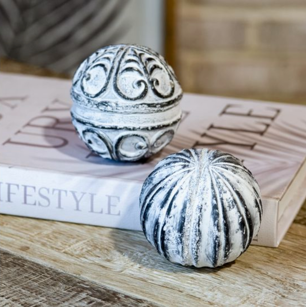 Two decorative hand-carved stone balls with a weathered finish, one with a whitewashed, dimpled texture and the other with an ornate pattern, placed on a wooden surface.