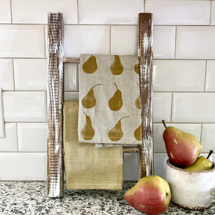 A set of two beige and gold tea towels with a pear motif, displayed on a wooden rack with pears and a white bowl in the background.