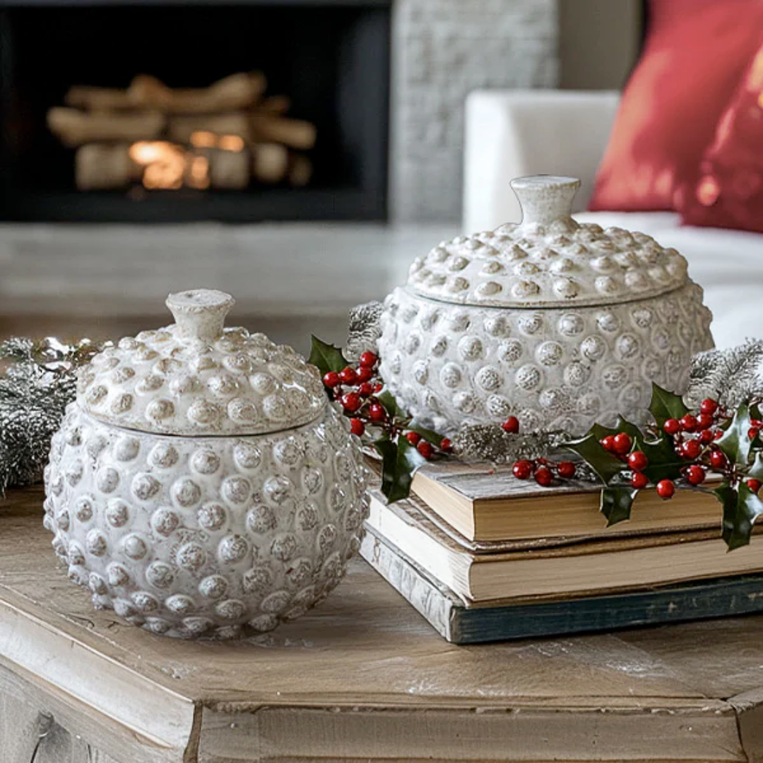 Two textured ceramic jars on a table with books and holly leaves, fireplace in the background