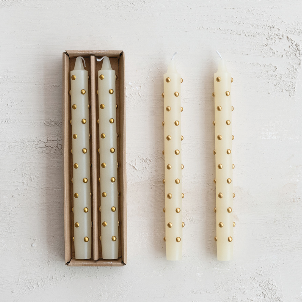 Two candles with gold studs next to their packaging on a light gray background