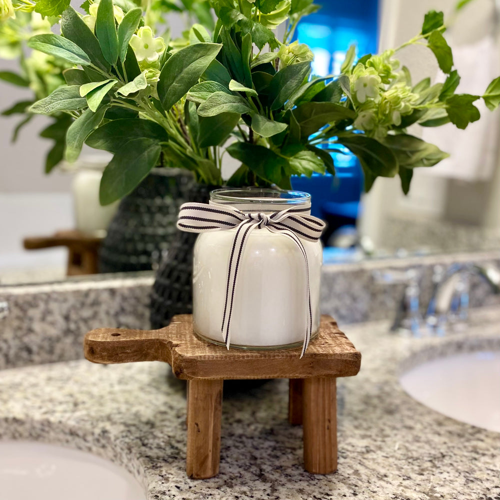 A vanilla caramel scented candle with a black and white striped ribbon on a wooden stand, set on a countertop with a plant in the background.