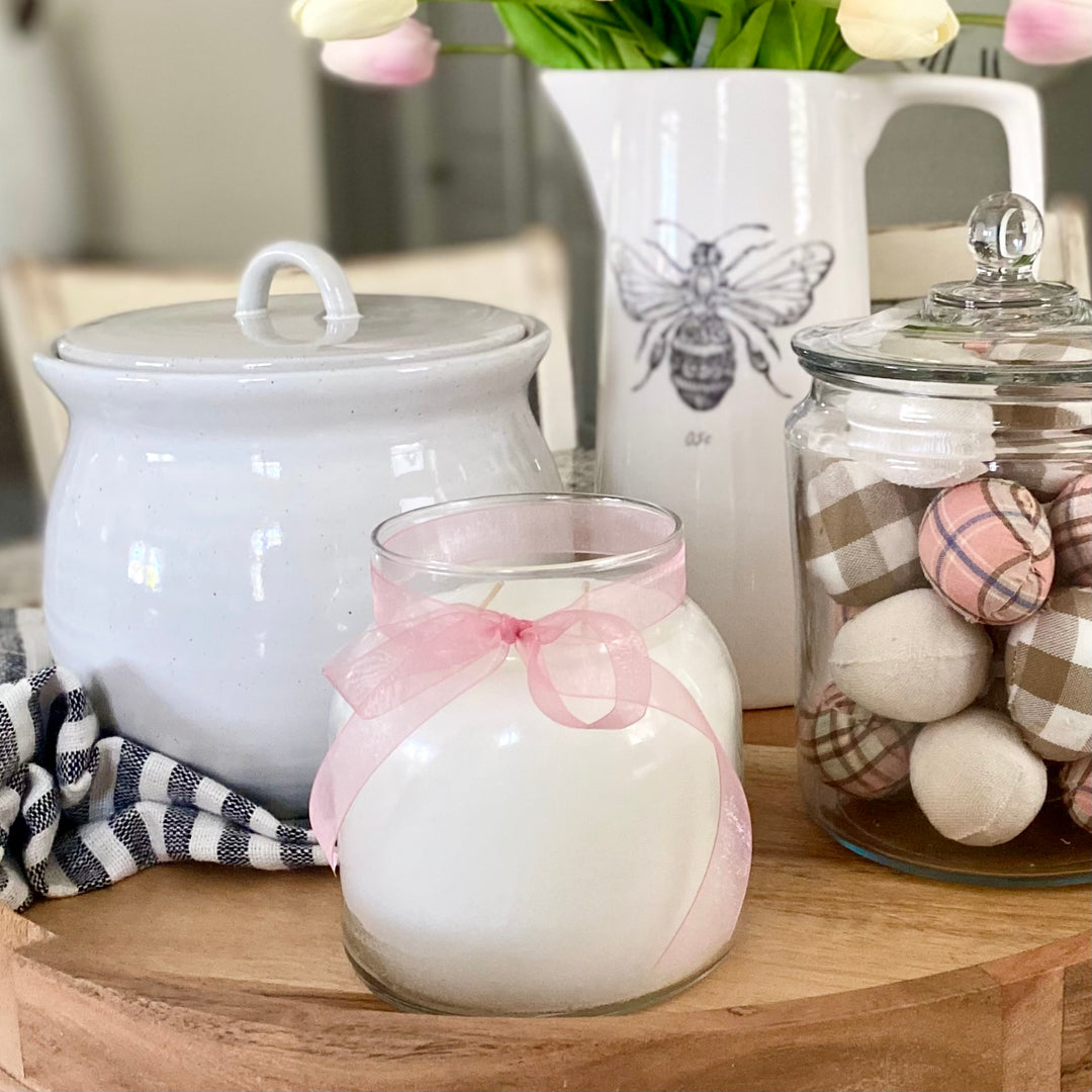 A vanilla scented white jar candle with a pink ribbon, alongside a black jar with a white lid featuring a bee design, and a glass jar filled with decorative eggs.