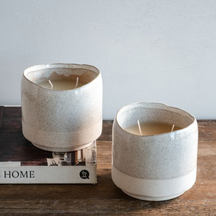 Two ceramic candles in rustic containers on a wooden surface with a book underneath.
